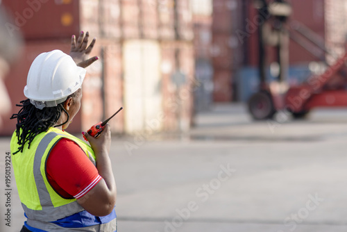 African woman engineer with safety vest and helmet holds worky talky, gives hand signals to stop forklift at container yard. Female foreman worker working at logistic shipping cargo container yard.