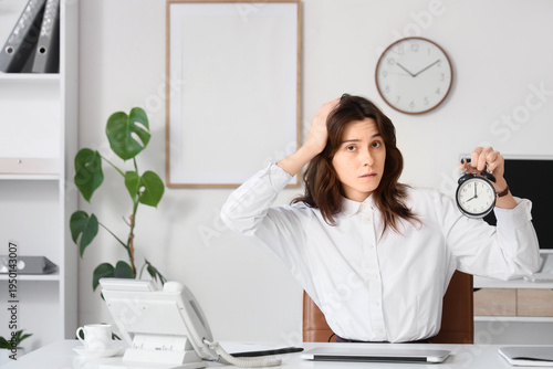 Confused young businesswoman with alarm clock at her workspace in office. Dea...