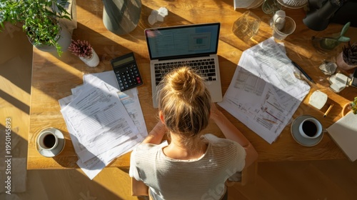 Top view of a young blonde woman holding her head in stress while sitting at a cluttered wooden table with documents, a calculator, and coffee.