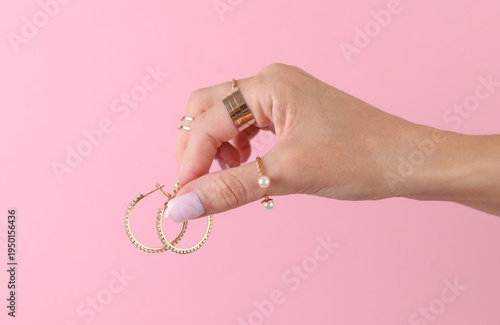 Woman's hand with many gold rings on her fingers holding earrings on pink background. Jewelry products