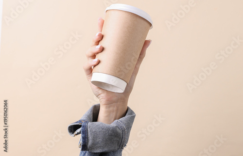 Female hand in denim jacket holds a disposable coffee cup on a beige background