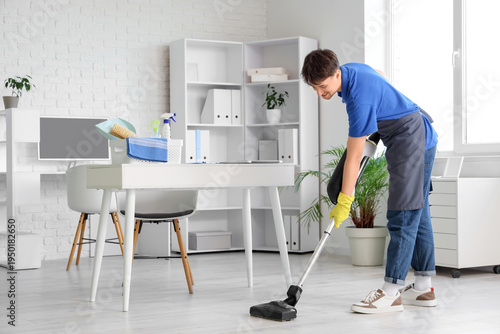 Young male janitor with modern vacuum cleaner cleaning floor in office