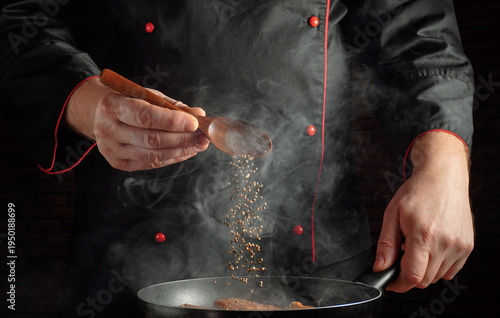 A chef in a black uniform uses a wooden spoon to sprinkle spices into a frying pan. Steam rises as food cooks on the stove. The setting is a busy restaurant kitchen at night