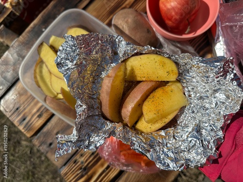 Sliced ​​potatoes with seasonings in baking foil near carrots on wooden table outdoors