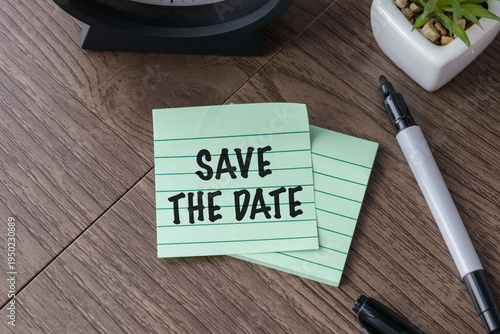 Conceptual image of a green sticky note with SAVE THE DATE text on a wooden desk, symbolizing event planning, appointment reminders, and deadline management.
