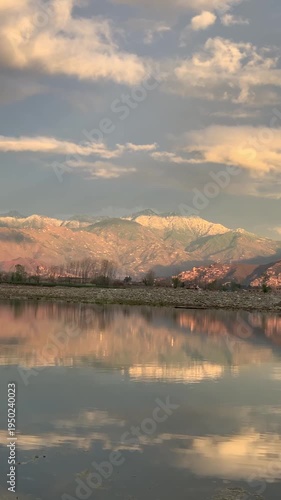 A scenic wide-angle landscape featuring a calm river in the foreground with a crisp mirror reflection of the surrounding environment. In the background, majestic mountains are bathed in the warm.