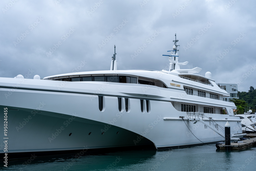 Fototapeta premium White luxury yacht floats calmly in harbor waters. Cloudy sky looms overhead, softening the scene. Modern design features sleek lines and large windows. Water reflects yacht's clean shape.