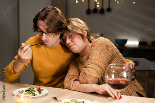 Young couple in love enjoying romantic dinner with wine at home husband eating while tired wife sleeps on his shoulder
