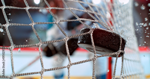 Close-up action shot of an ice hockey puck hitting the goal net during a scoring moment. Dynamic sports scene with ice spray and a blurred hockey player in the background.