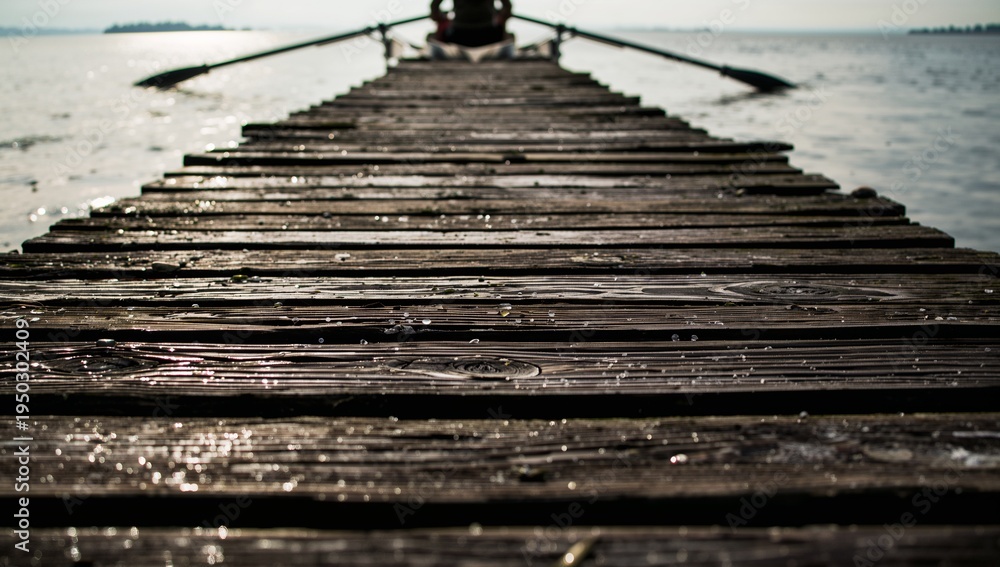 Obraz premium Leading wooden pier stretching to rowboat at lakeshore with person wearing dark coat, oars glinting