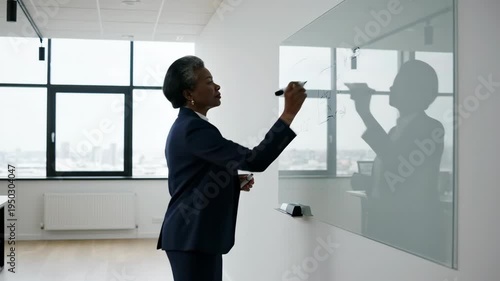 african american senior businesswoman writing on glass whiteboard in modern office. corporate strategy planning, brainstorming session, leadership concept.