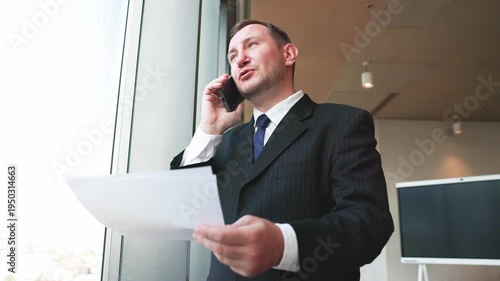 Professional businessman speaking on smartphone while holding financial report near office window during work discussion.