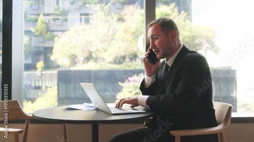 Professional businessman speaking on smartphone while holding financial report near office window during work discussion.