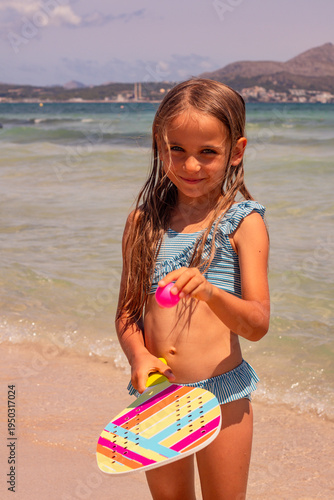 Young girl with long hair wearing a striped bikini holds a colorful paddle and ball while standing on a sandy beach near the water's edge