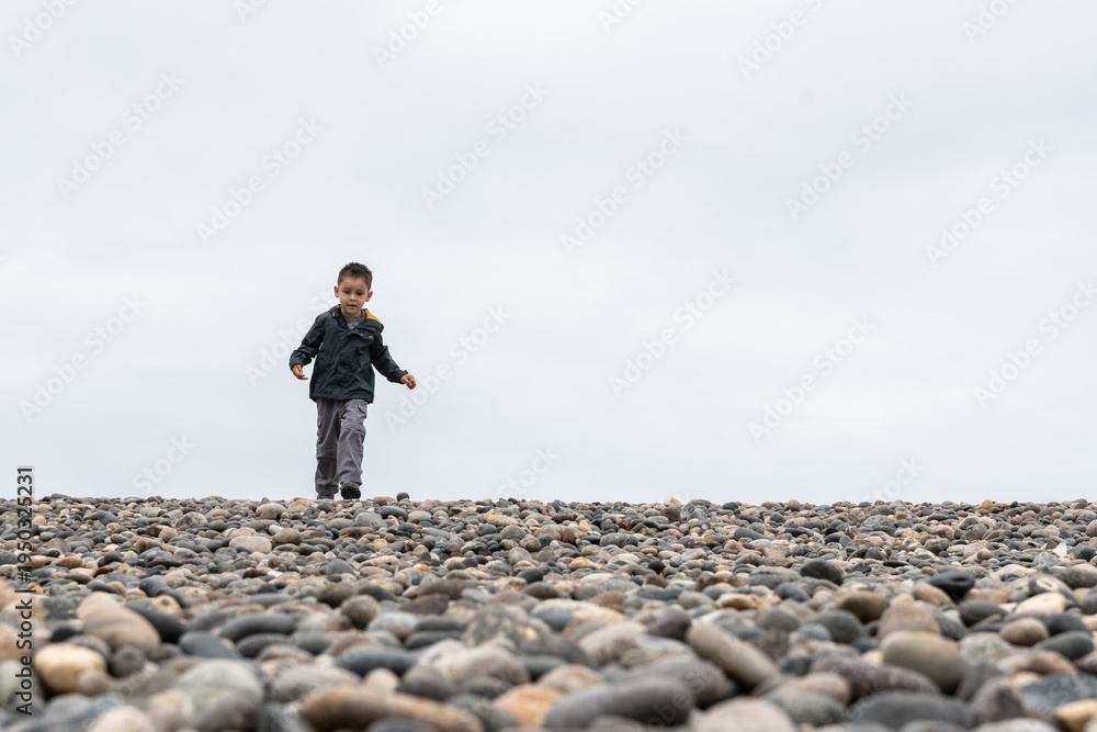 Obraz premium Little boy walking on a rocky beach under cloudy sky with copy space