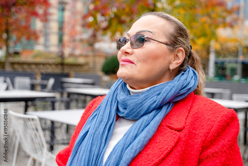 Woman wearing sunglasses and a red coat looks up while walking in a park during autumn