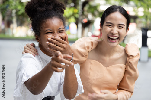 Two young women, Asian and African friends having a good time, talking and laughing together, pointing at you