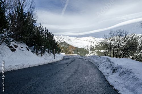 A wide-angle, low-perspective shot of a clean asphalt road curving through a winter mountain landscape. The road is flanked by snow banks and pine trees