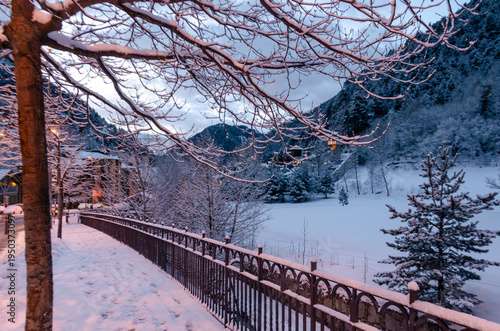 A serene twilight landscape in a winter mountain village. In the foreground, snow-dusted bare branches frame the view, while a decorative metal fence leads the eye toward a snow-blanketed field.
