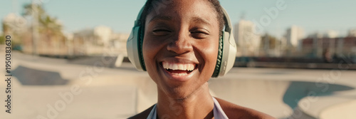 A girl in headphones stands in the background of a skatepark and laughing. Panoramic