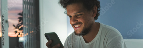 Guy is sitting on the bed in the bedroom, watching video, movie on mobile phone. Panoramic
