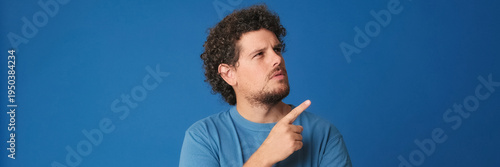 Guy with curly hair dressed in blue t-shirt is thinking over difficult choice, imagining plan in his mind, having an idea, isolated on blue background in the studio. Panoramic