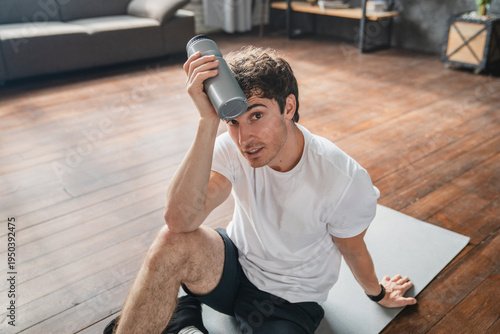 Man drinking water from bottle after yoga workout at home, resting in living room and hydrating body. Fitness routine, recovery, wellness and healthy lifestyle, training, refresh concept.