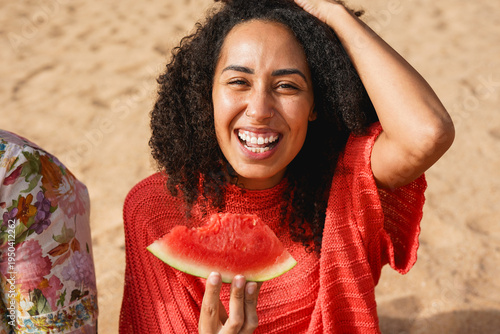 Happy young african woman smiling on camera while eating watermelon on the beach - Food, healthy lifestyle and summer vacation concept
