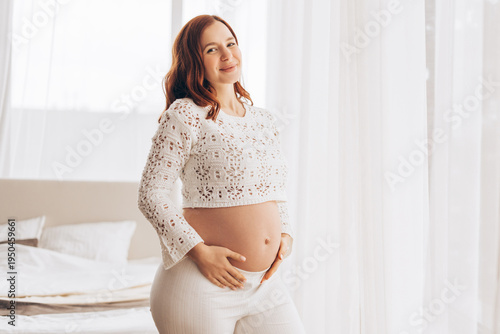 Free Download Smiling red-haired pregnant young woman touching belly near window and looking at camera - GFXLoad