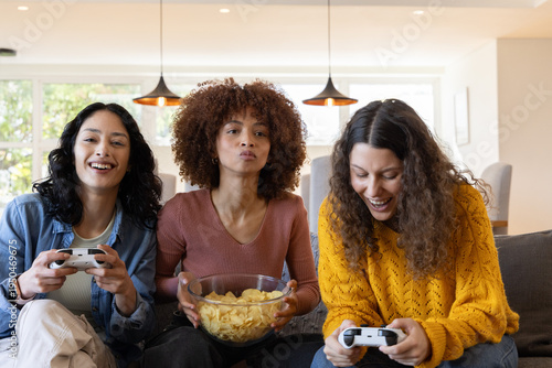 Diverse female friends sitting on gray sofa holding controllers and bowl of chips in living room