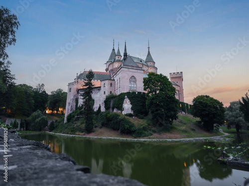 Atmospheric blue hour view of Bojnice Castle mirrored in calm moat water, surrounded by trees and evening light in Bojnice, Slovakia.