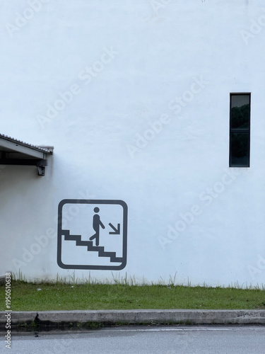 An emergency exit sign showing a figure descending stairs, affixed on a white wall with a small rectangular window. 