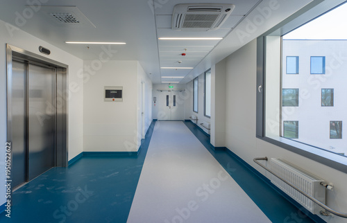 Modern hospital corridor interior with elevators and emergency exit signs.