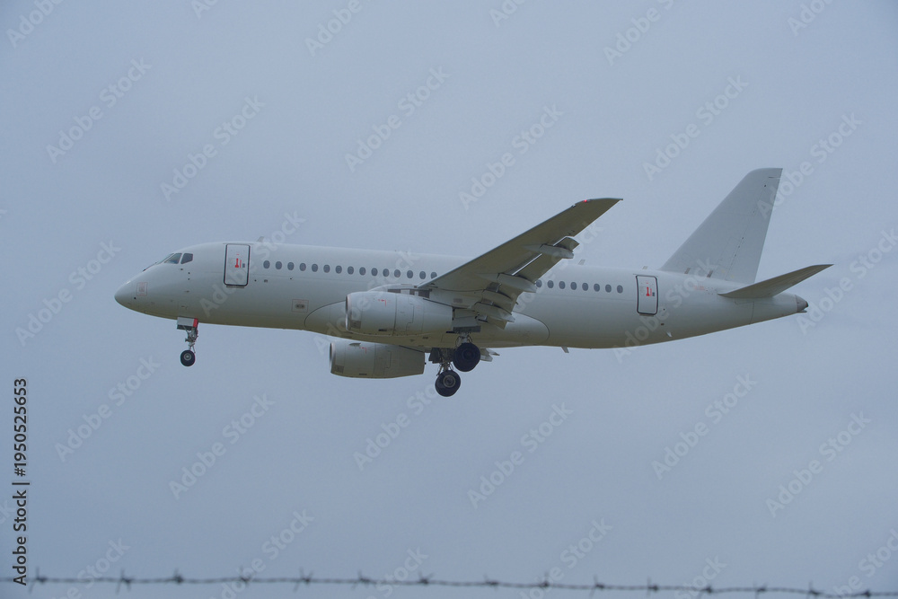 Obraz premium White Plane Approaching Runway Behind Barbed Wire Fence. White Commercial Airplane Approaching Runway Behind Barbed Wire Fence on Overcast Day