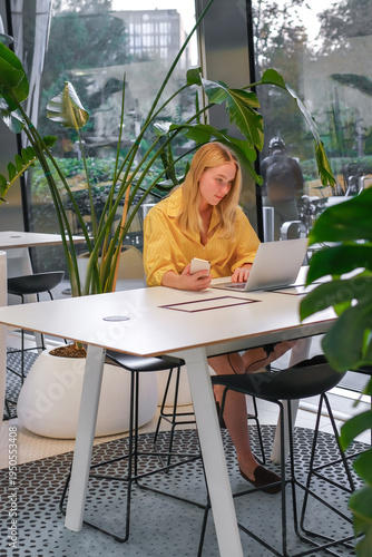 Gen Z remote worker in coworking space, young woman using laptop at modern shared desk with indoor plants, focused productivity, hybrid work, work-life balance concept