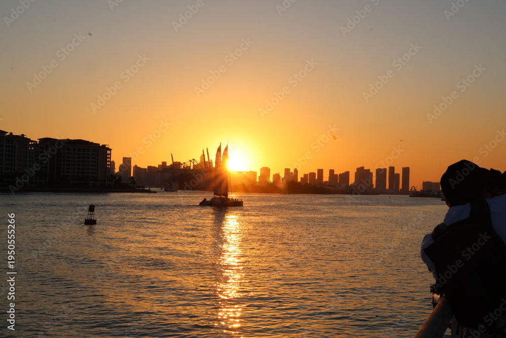 Fototapeta premium Silhouette of the mainland Miami skyline with cranes and construction sites along the canal at South Pointe Park during sunset, showing urban development and waterfront scenery.