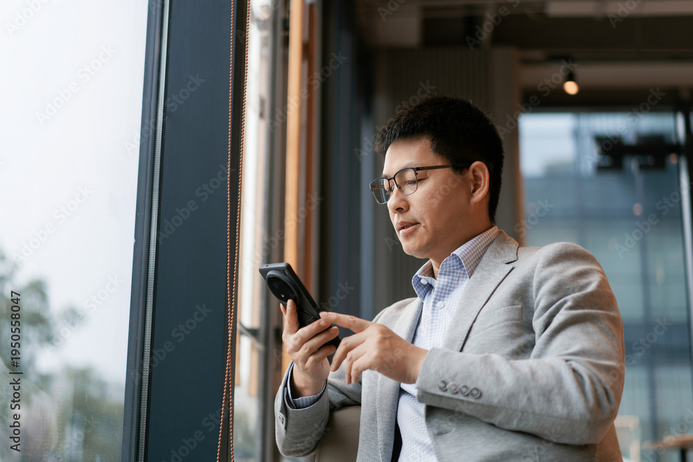 Fototapeta premium A bespectacled businessman in a suit checks his smartphone while standing by the window in a modern office