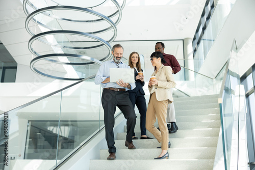 Diverse team of professionals walking down a modern office staircase, collaborating over a laptop and discussing a project while smiling