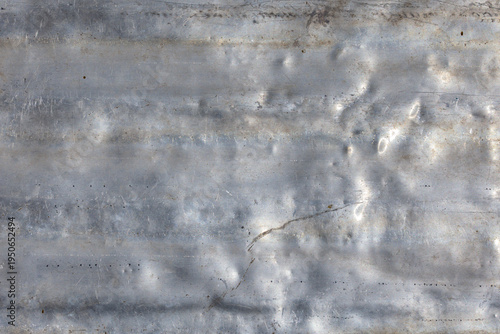 Rough aluminum sheet displays textured surface with scratches and damage. Outdoor industrial setting highlights weathered appearance under bright daylight.