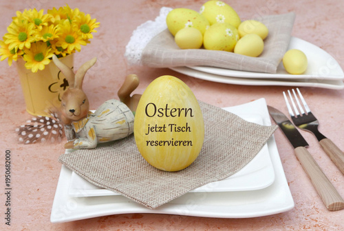 Eine harmonische Tischdekoration mit einem gelben Osterei, auf dem „Ostern jetzt Tisch reservieren” steht, weißen Tellern, Besteck und einer liegenden Hasenfigur vor einer Vase mit gelben Blumen.