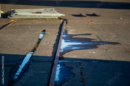 Old rusty railway tracks on cracked asphalt with water puddles and blue sky reflection in industrial area