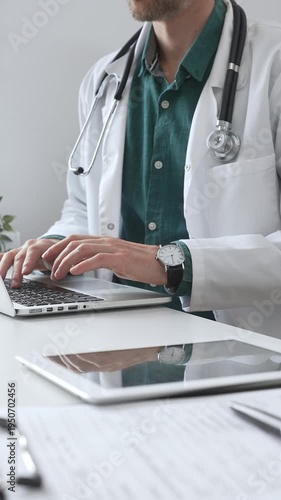 Doctor man wearing a white coat over green shirt, and stethoscope is typing on a laptop in a bright medical office, showcasing the intersection of healthcare and technology. Medicine and health care