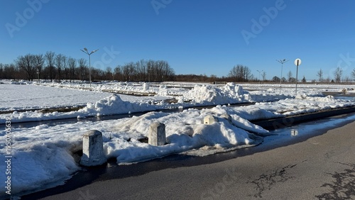 Foto Melting snow piles and puddles on a wet asphalt parking lot bordered by concrete posts on a sunny early spring or late winter day under a clear blue sky