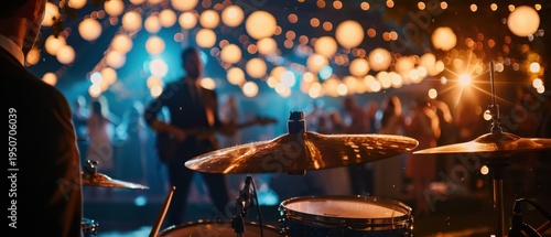 Close-up of a drum kit cymbal in focus during a live band performance. Blurred background of musicians, a dancing crowd, and warm glowing string lights bokeh. Outdoor concert or wedding party concept.