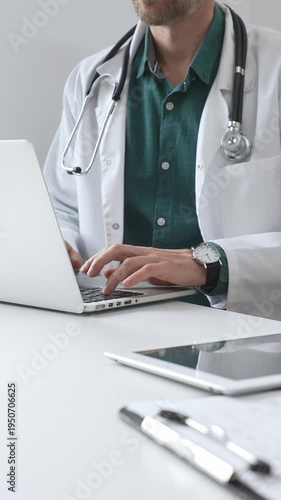 Doctor man wearing a white coat over green shirt, and stethoscope is typing on a laptop in a bright medical office, showcasing the intersection of healthcare and technology. Medicine and health care