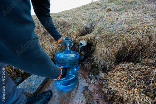 Fotografi Gloved hands hold a blue jug under a short white pipe as clear spring water flows from a rural hillside, with coarse yellow brown grass and stones in muted late light