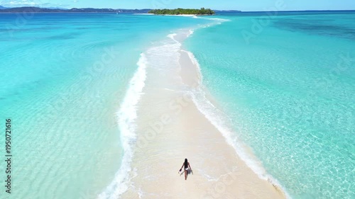 Aerial drone view of a woman walking along a narrow white sandbar between shallow turquoise waters, with a small island visible in the distance in Madagascar