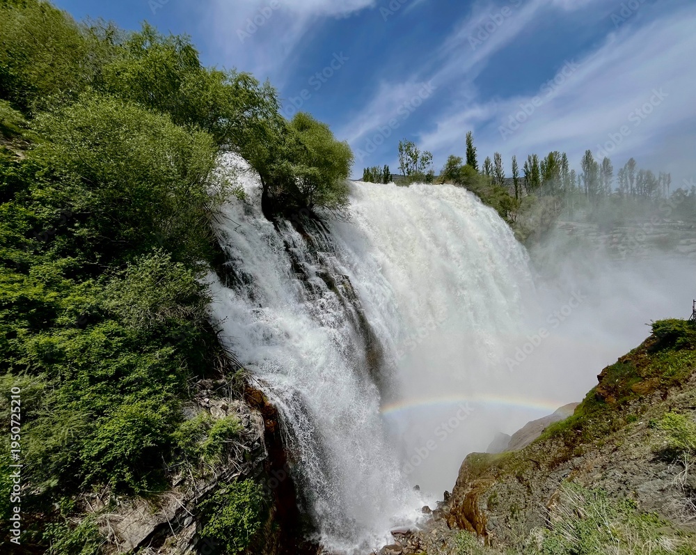 Fototapeta premium Breathtaking view of a powerful majestic waterfall cascading down rocky cliffs with a beautiful rainbow in the mist.