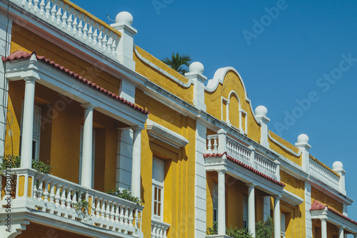A vibrant yellow colonial building in Cartagena under a clear blue sky, featuring ornate white balconies, classic columns, and decorative roofline balustrades.