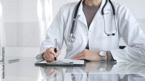 Quadro su tela Female doctor in white coat with stethoscope writing patient notes on clipboard, completing medical records in clinic office